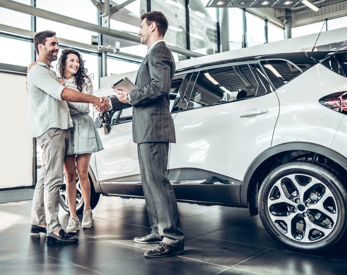 A smiling couple shakes hands with a salesman in a suit, standing beside a shiny white SUV in a bright showroom.