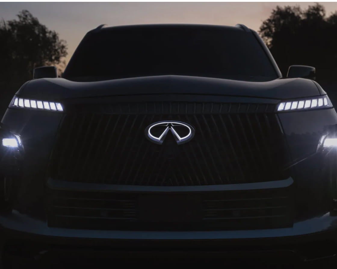 Front view of an Infiniti SUV at dusk, with headlights and logo illuminated, creating a sleek and modern appearance against a shadowy backdrop.