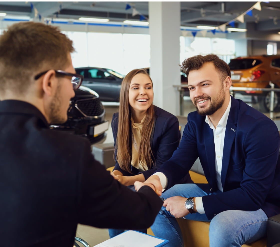 A couple in a car dealership shakes hands with a salesperson. They are smiling, indicating a successful deal.