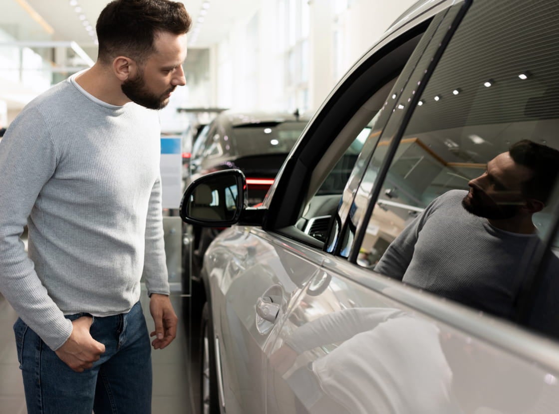 A man in a gray sweater inspects a car at a dealership, reflecting thoughtfully in the vehicle's window.