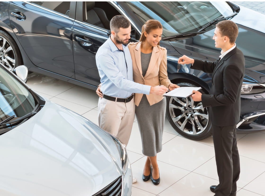 A couple stands in a car showroom, reviewing a document with a salesman handing over car keys.